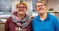 Zwei Frauen mit Brillen lächeln in einem Innenraum mit Fenstern im Hintergrund