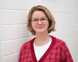 Frau mit blonden Haaren und Brille vor einer weißen Wand.